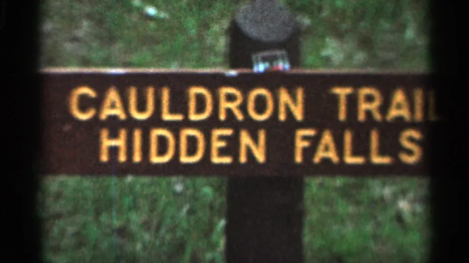 1966 Hamel, Minnesota 8mm frame of a wooden Cauldron Trail Hidden Falls sign in a shaded wooded setting.