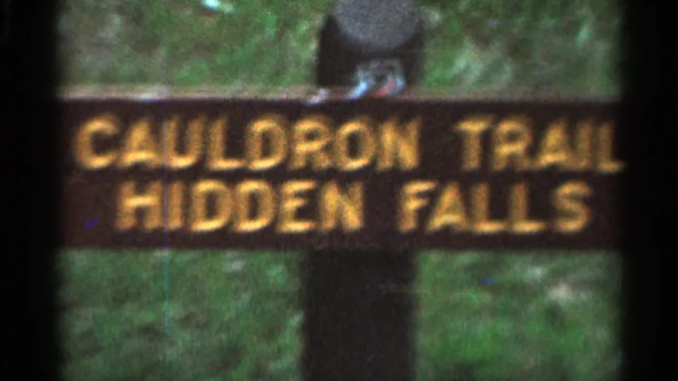 Soft 8mm view of the Cauldron Trail Hidden Falls sign, with yellow letters and green woods behind it.