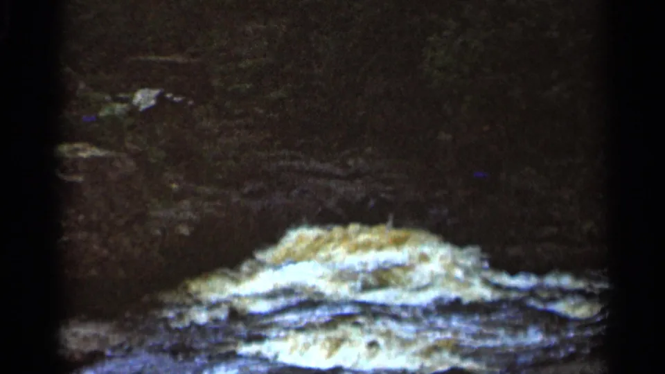 1966 8mm frame from Hamel, Minnesota showing white water spreading over layered rock in a shadowed natural area.