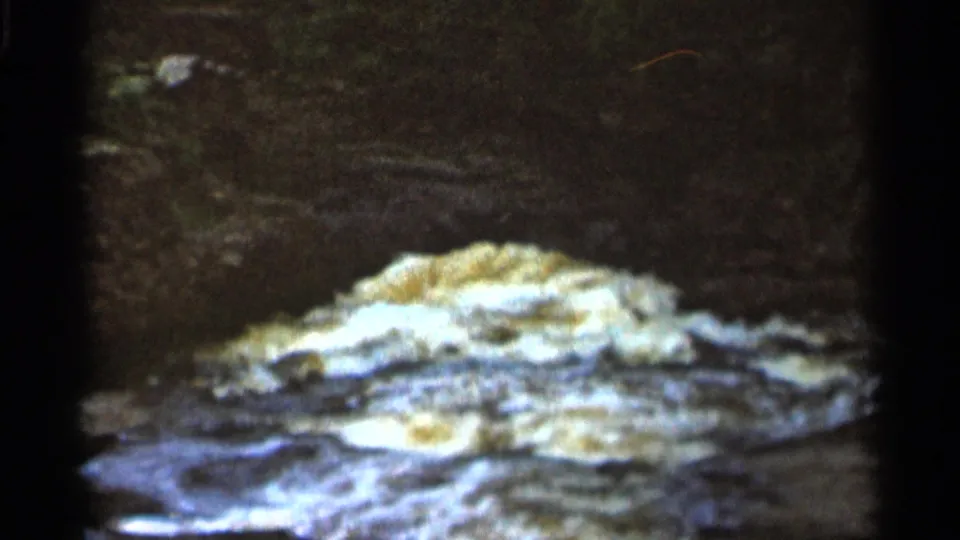 1966 8mm Hamel, Minnesota frame showing low frothy rapids crossing rock with shadowed stone in the background.