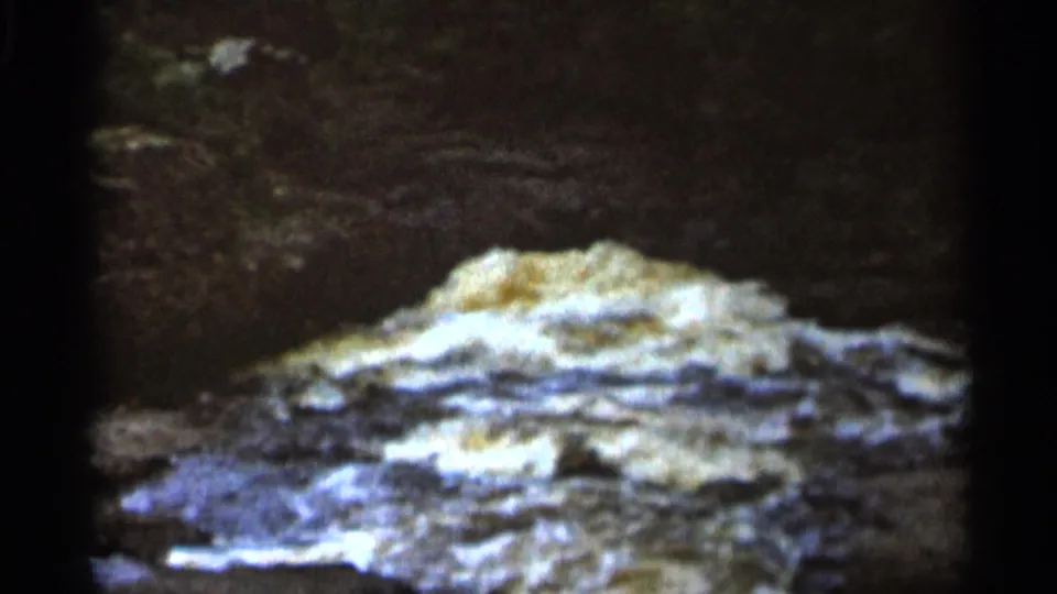 1966 8mm frame from Hamel, Minnesota showing frothy stream water moving over rocks below a dark natural backdrop.