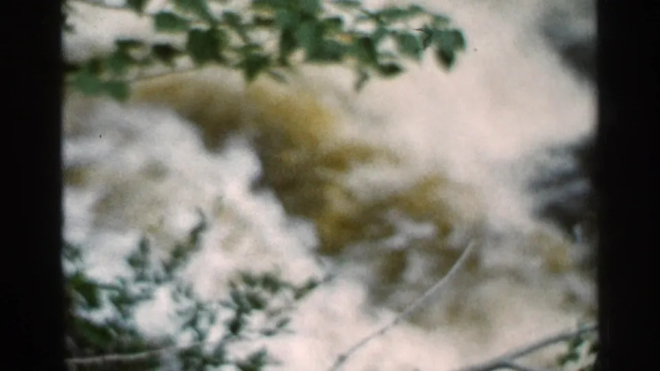 1966 Hamel, Minnesota 8mm frame with green leaves in front of foamy muddy rapids and a blurred shoreline view.