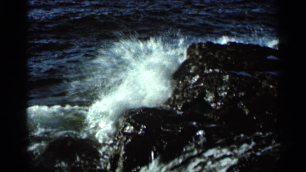 Wave spray bursts against dark shoreline rocks in daylight.