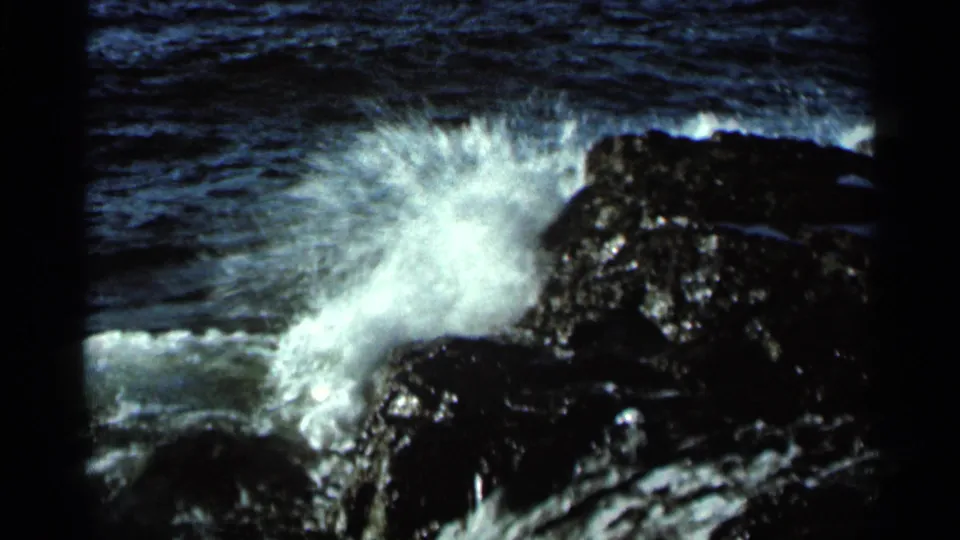 1966 8mm frame of white surf breaking over dark coastal rocks, with choppy blue water behind it.