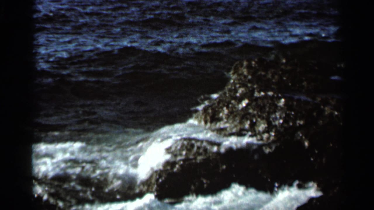 Foamy surf wraps around a dark rock outcrop near open water.