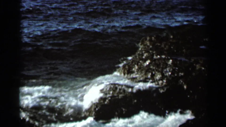 1966 8mm frame showing white foam curling around dark shoreline rocks under daylight.