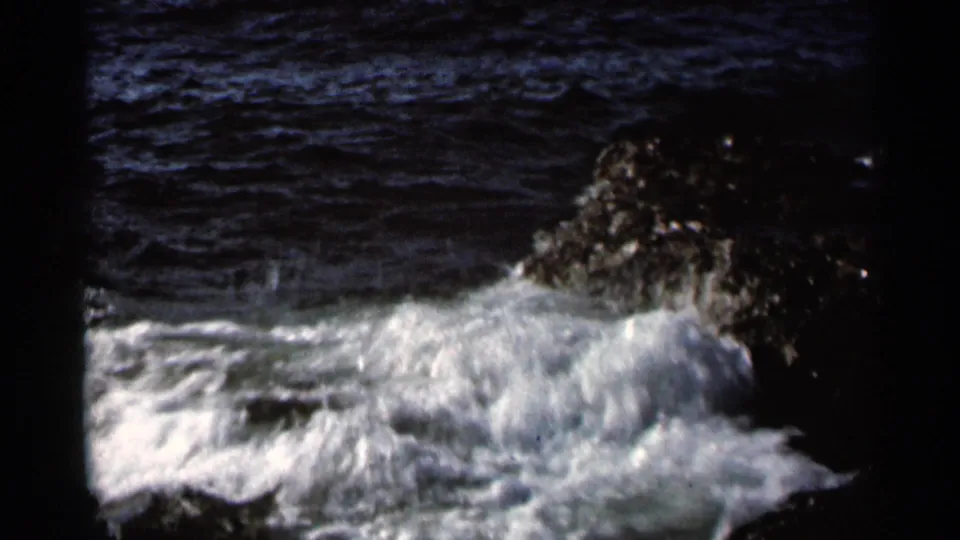 1966 8mm frame of white water spreading over dark rocks, with textured blue sea in the background.