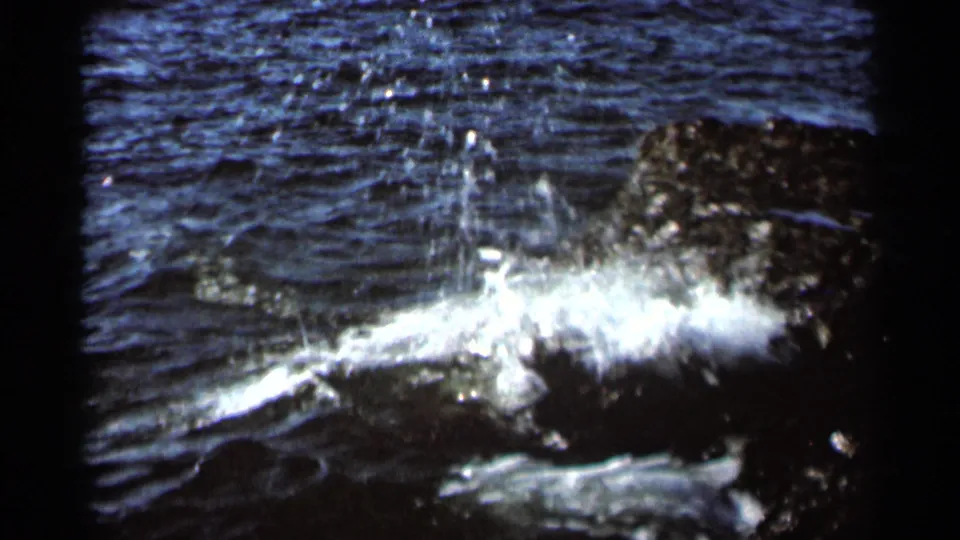 1966 8mm frame of surf striking dark shoreline rocks, with spray rising against rippled water.