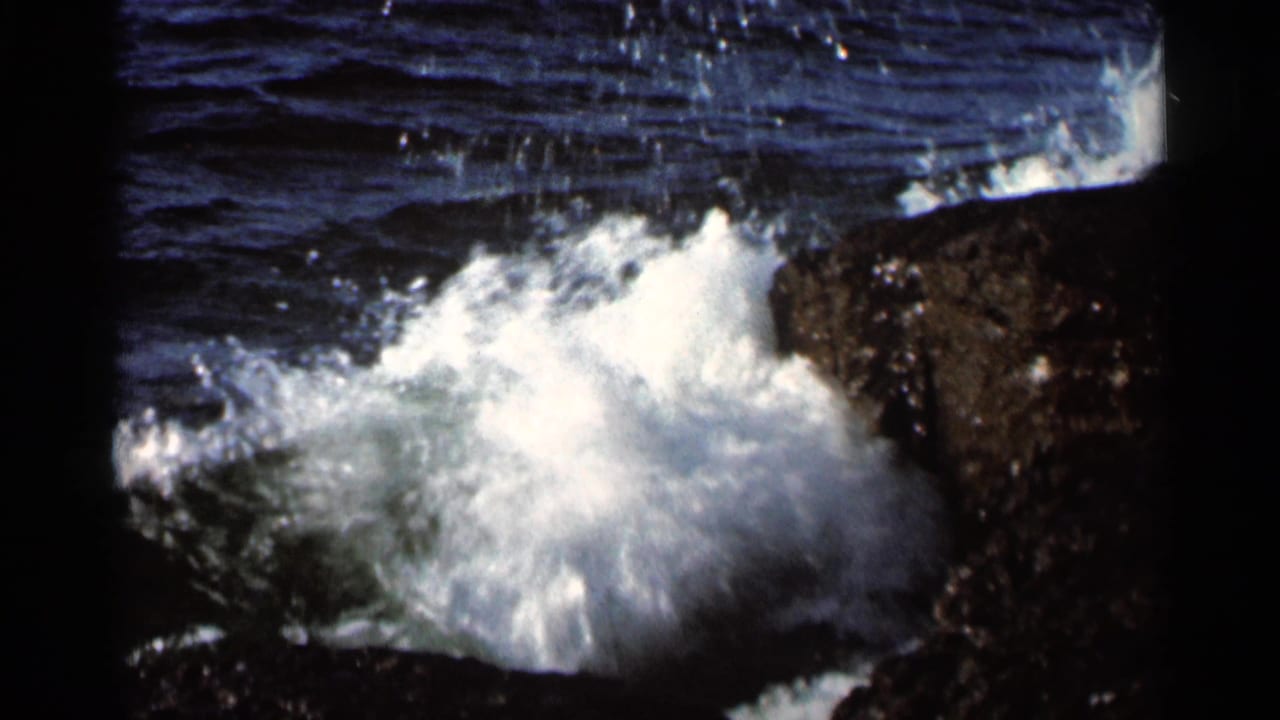 1966 8mm frame showing white waves breaking hard against dark rock along a rugged shoreline, with deep blue water above.
