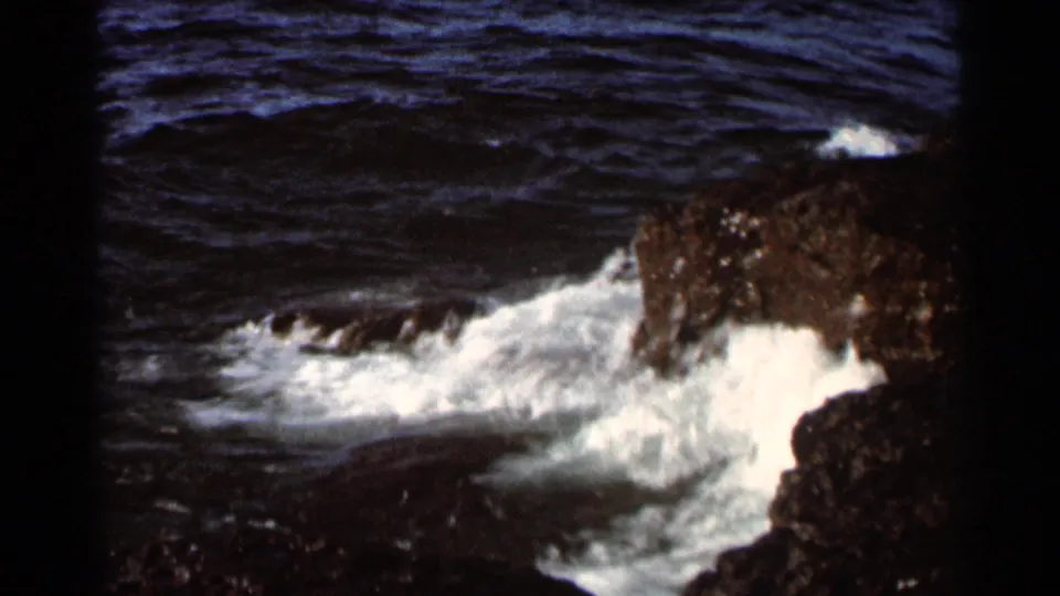 1966 8mm frame showing waves and foam moving into a dark rocky inlet, with rough water and jagged stone edges visible.