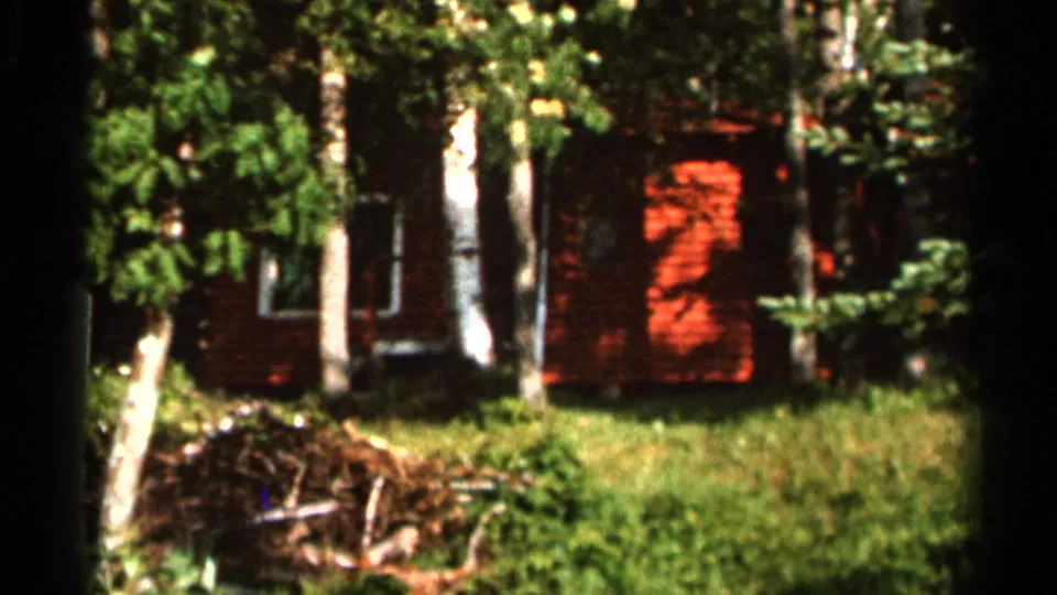 1966 8mm frame showing a red cabin among trees, with bright sunlight on the wall, green grass below, and a brush pile in front.