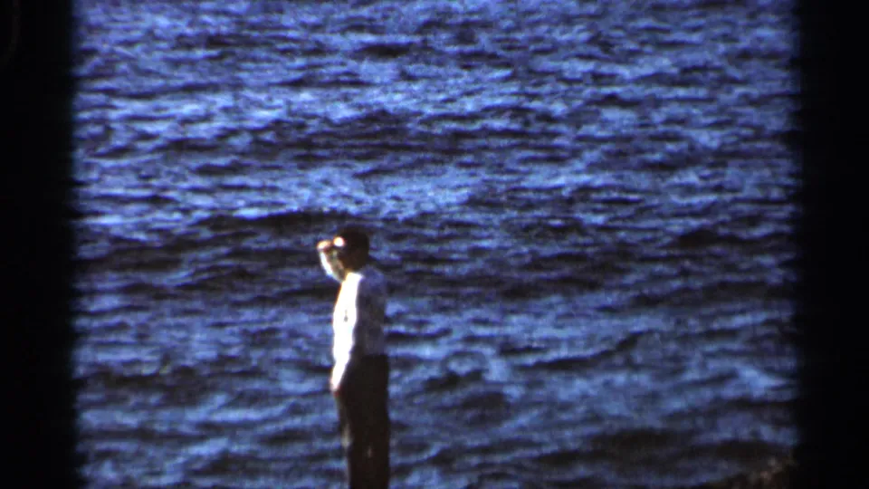 1966 8mm frame showing a distant adult man in profile at the edge of a large, choppy body of water.