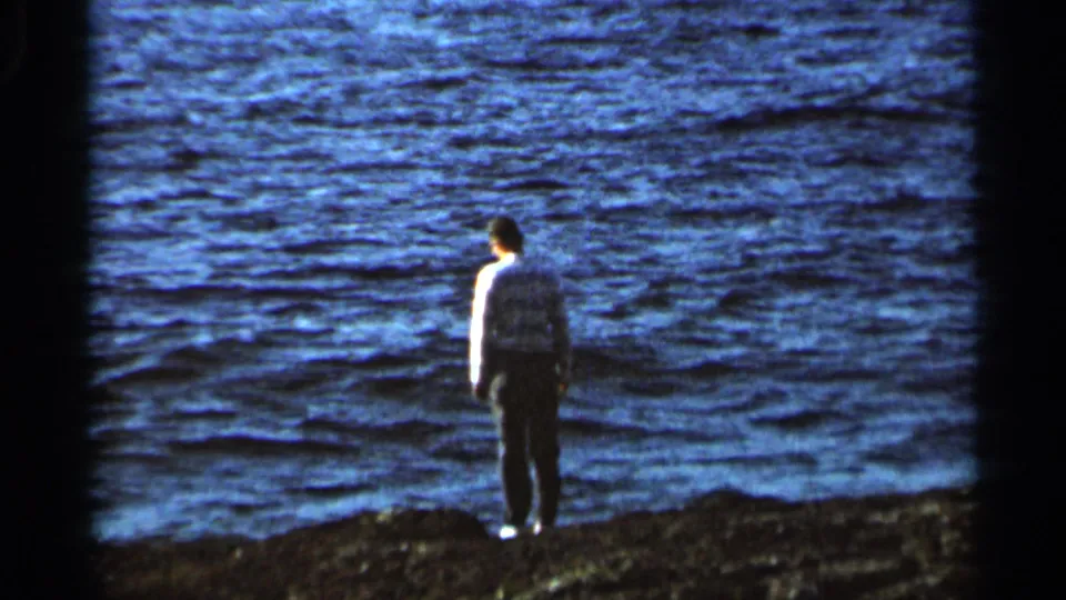 Adult man stands with his back turned on a rocky shoreline, looking out across rough blue water in 1966 home-movie footage.