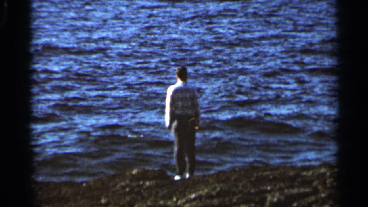 In 1966 Hamel, Minnesota home-movie footage, a man in a light shirt and gray trousers stands on a shoreline facing blue water.