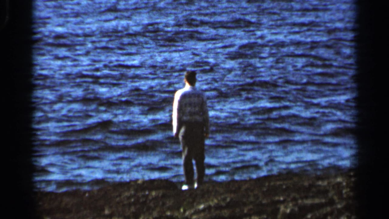 Man stands near the edge of rippling water with his back to the camera.