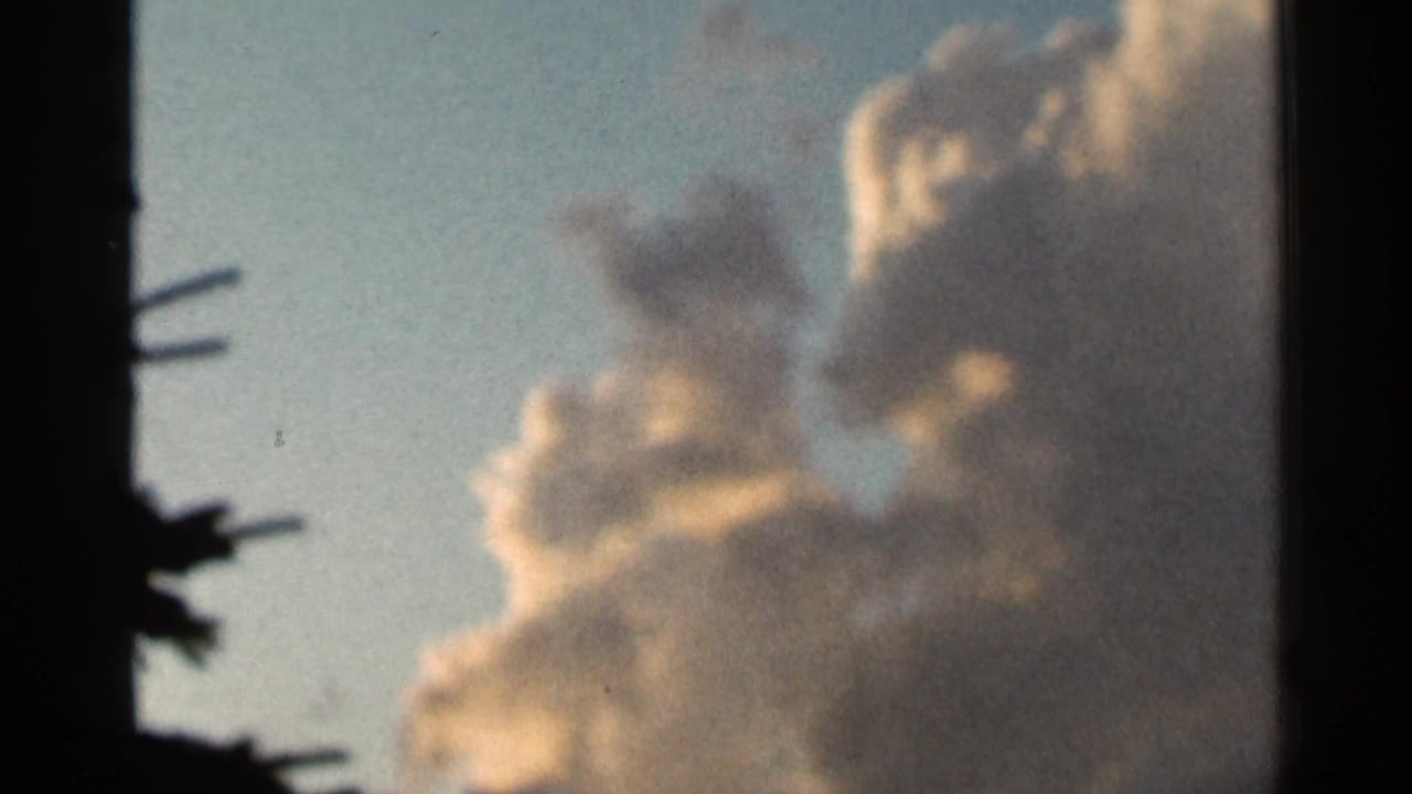 1966 Hamel, Minnesota 8mm frame showing pale yellow-white clouds against blue sky, bordered by dark tree shapes at the left.