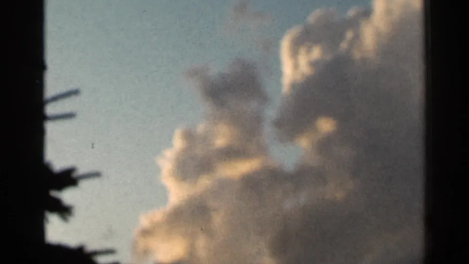 1966 Hamel, Minnesota 8mm frame showing pale yellow-white clouds against blue sky, bordered by dark tree shapes at the left.