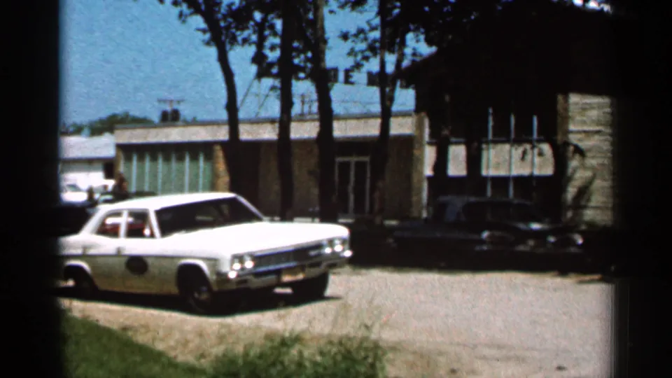 Wide 1966 Hamel, Minnesota exterior with a white sedan, gravel lot, trees, large windows, and a low business building.