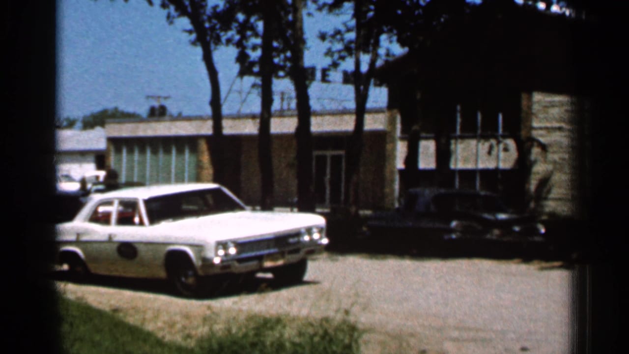 1966 Hamel, Minnesota 8mm frame showing a white sedan on a gravel driveway near a low building with trees casting shade.
