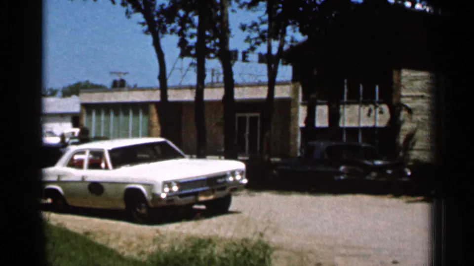1966 Hamel, Minnesota 8mm frame showing a white sedan on a gravel driveway near a low building with trees casting shade.