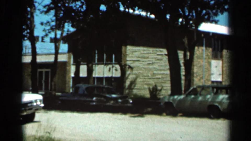 1966 Hamel, Minnesota frame showing the end of the pan across a stone-faced building, trees, a dark parked car, and partial vehicles.