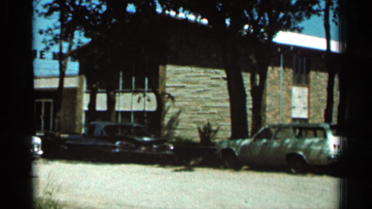 1966 Hamel, Minnesota frame showing a light station wagon, darker cars, tree shadows, and a brown stone building exterior.