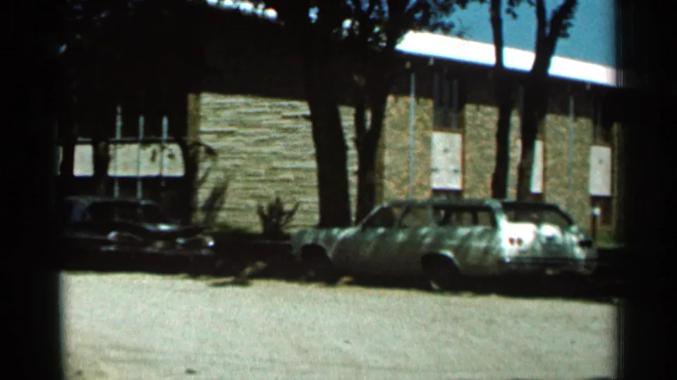 1966 Hamel, Minnesota view of parked cars beside a brown stone building, partly shaded by tall roadside trees.