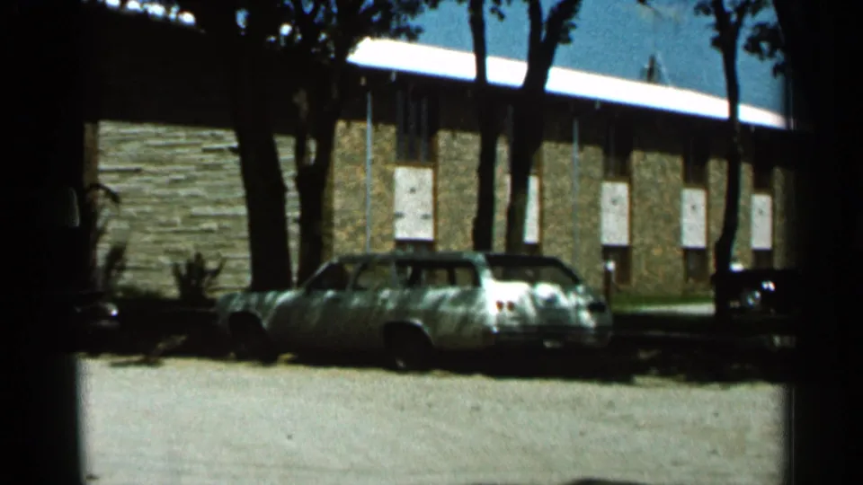 1966 Hamel, Minnesota home-movie frame showing a brick low-rise building with parked cars, tree trunks, and bright daylight.