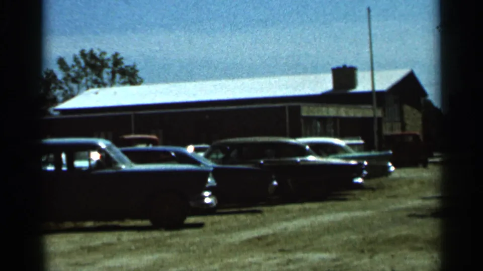1966 Hamel, Minnesota home-movie still with parked cars, a brick building, gravel ground, and trees in the background.