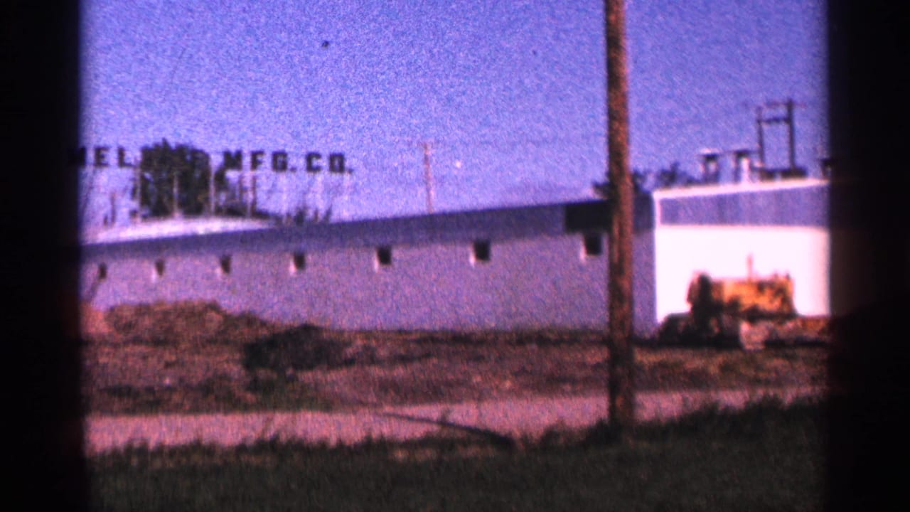 White industrial warehouse with rooftop company sign, utility pole, dirt lot, and blue sky.