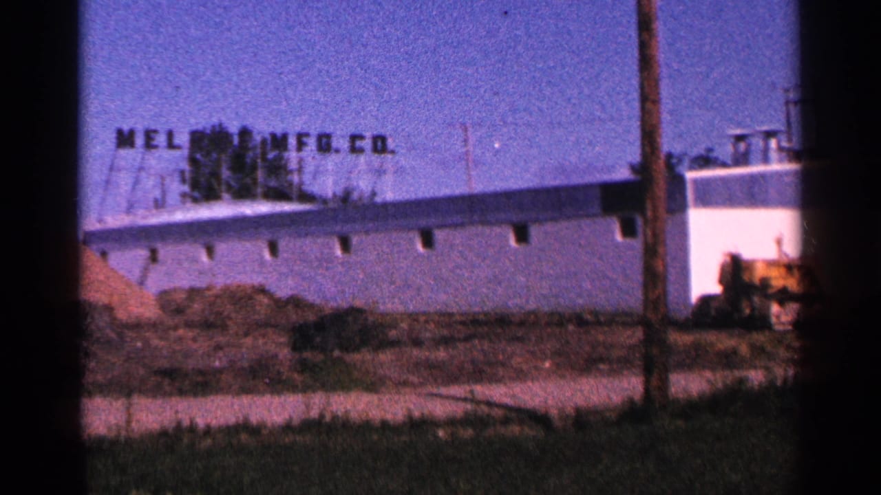 Industrial building with rooftop sign, pole in foreground, dirt piles, and yellow equipment at right.