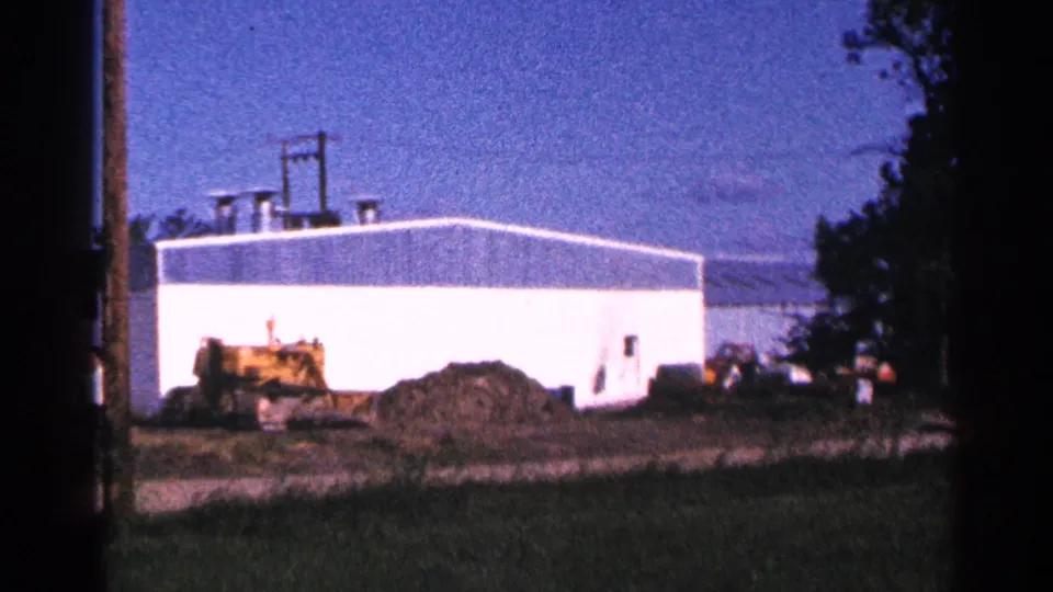 1966 Hamel, Minnesota 8mm frame showing the white warehouse end, yellow bulldozer, dirt pile, grass, and trees along the edge.