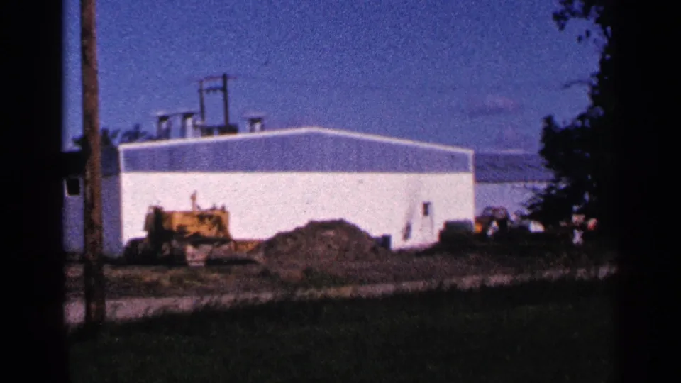 1966 Hamel, Minnesota 8mm frame showing a white industrial building under construction with a yellow machine and dirt pile in front.