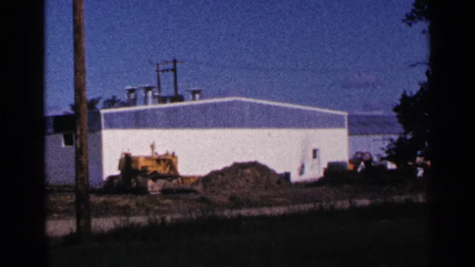 1966 home-movie view of a white industrial building in Hamel, Minnesota, with a yellow machine and earthwork in front.