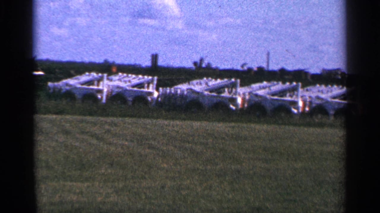 1966 Hamel, Minnesota home-movie frame showing white farm equipment or trailers in a green field beneath a pale blue sky.