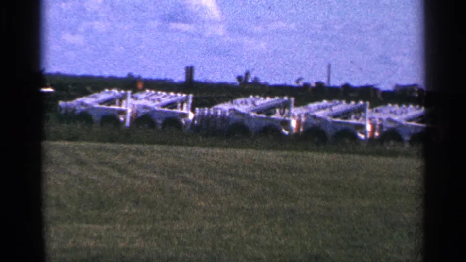 1966 Hamel, Minnesota home-movie frame showing white farm equipment or trailers in a green field beneath a pale blue sky.