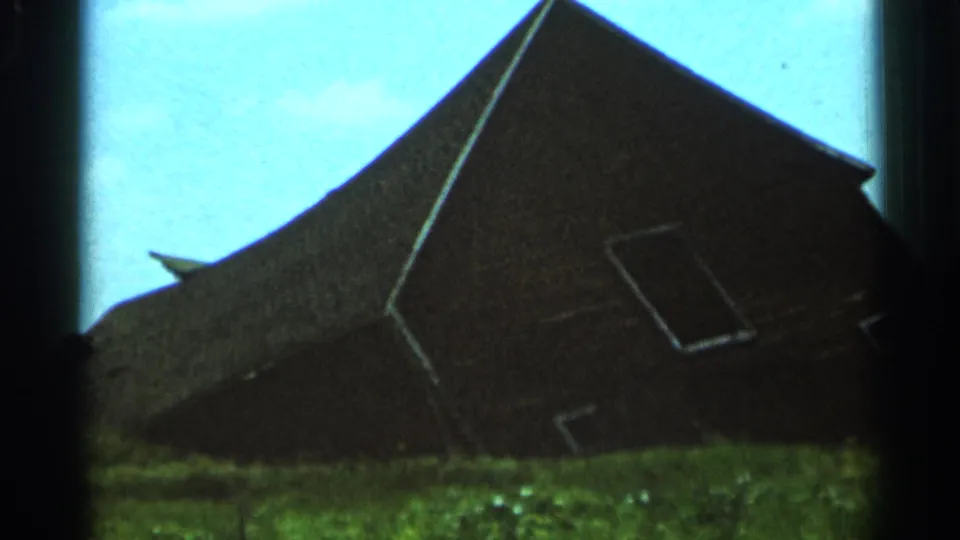 1966 Hamel, Minnesota frame of a steep-roofed dark barn, partially silhouetted against bright blue sky and grass.
