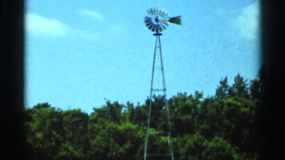 1966 Hamel, Minnesota 8mm frame with a tall metal windmill above dense trees and a bright blue sky.