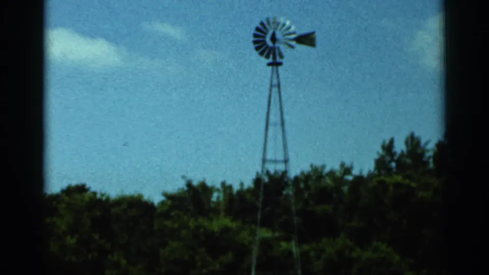 1966 Hamel, Minnesota 8mm frame showing a centered farm windmill, leafy treeline, pale cloud, and soft film grain.