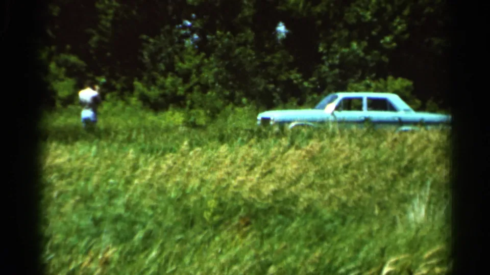 1966 Hamel, Minnesota 8mm frame showing a distant person in a field beside a light blue car near trees.
