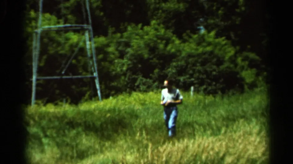 The man walks toward the camera through tall grass, holding a camera or object, with a metal power line tower behind him.