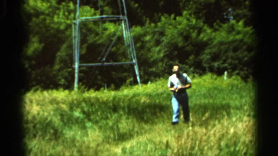 A man in a light shirt and blue jeans stands in the grassy Hamel field with a metal tower and trees behind him.
