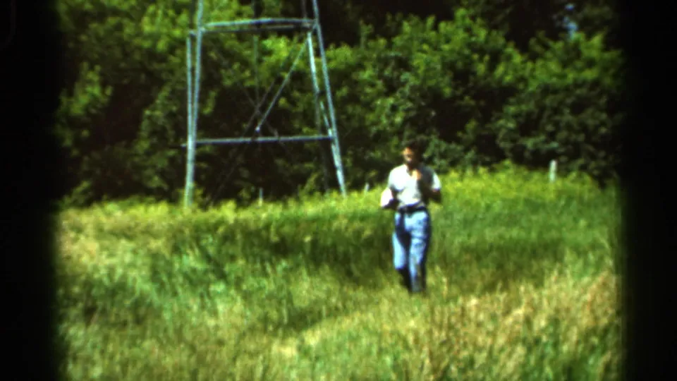 1966 8mm frame from Hamel, Minnesota, showing a man in a light shirt and jeans walking through tall grass near a metal utility tower.