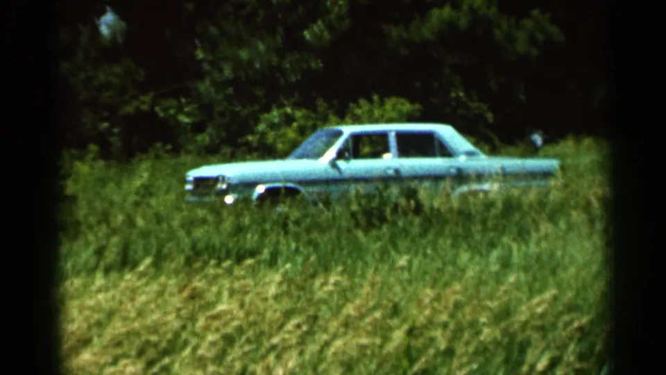 1966 rural Hamel, Minnesota frame showing a turquoise sedan parked in tall grass, framed by soft 8mm color and dark edges.