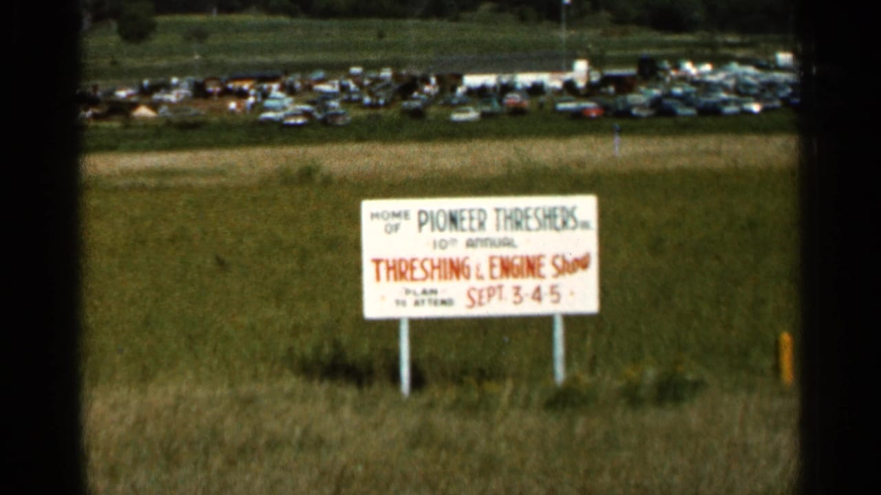 White Threshing & Engine Show sign in a field with parked cars beyond.