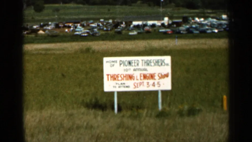 A white sign for the 10th Annual Threshing & Engine Show stands in a grassy field near a distant parking area in Hamel, Minnesota.