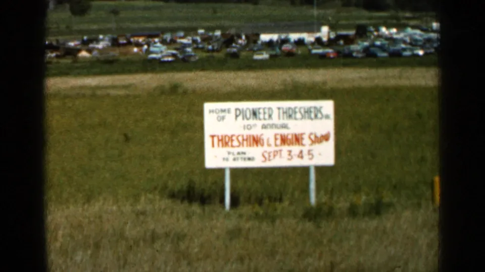 The Pioneer Threshers event sign is framed across grass, with rows of 1960s-era cars visible in the background.