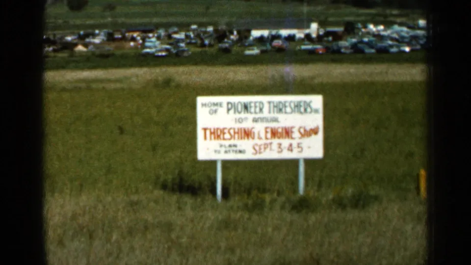 A rural show sign for the Threshing & Engine Show stands in tall grass, with many parked cars beyond the field.