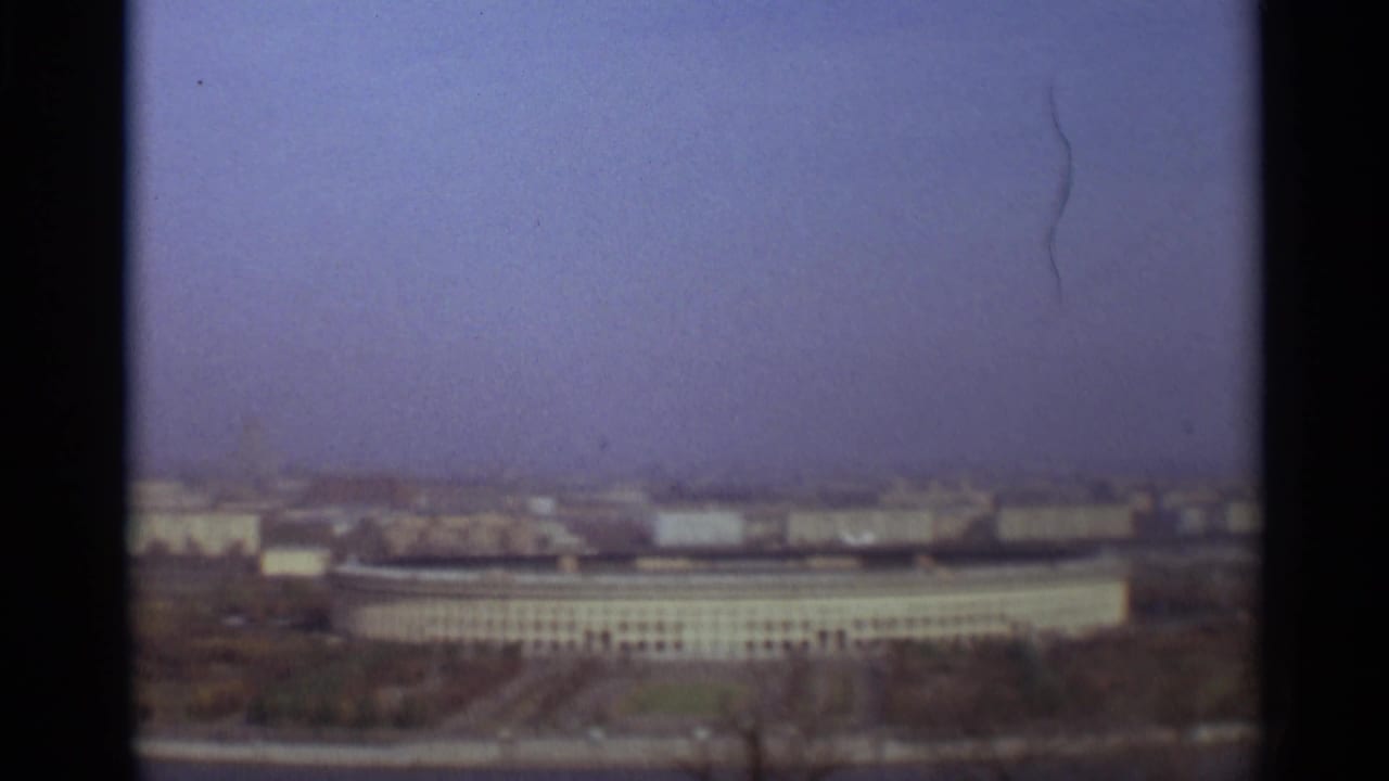 Hazy elevated view of large low buildings and distant city skyline behind dark film borders.