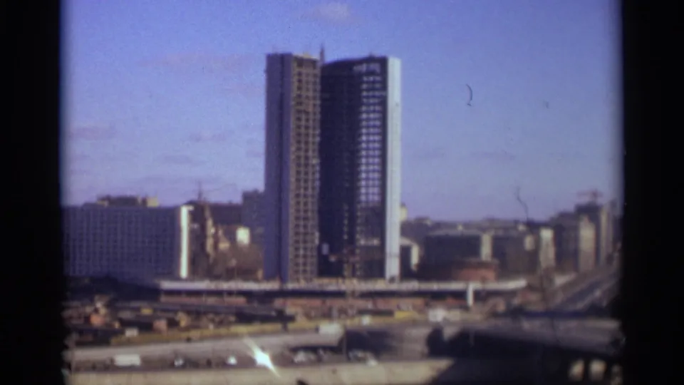 1966 Moscow skyline view with a tall high-rise, nearby cranes, wide roads, and distant urban buildings in grainy 8mm color.